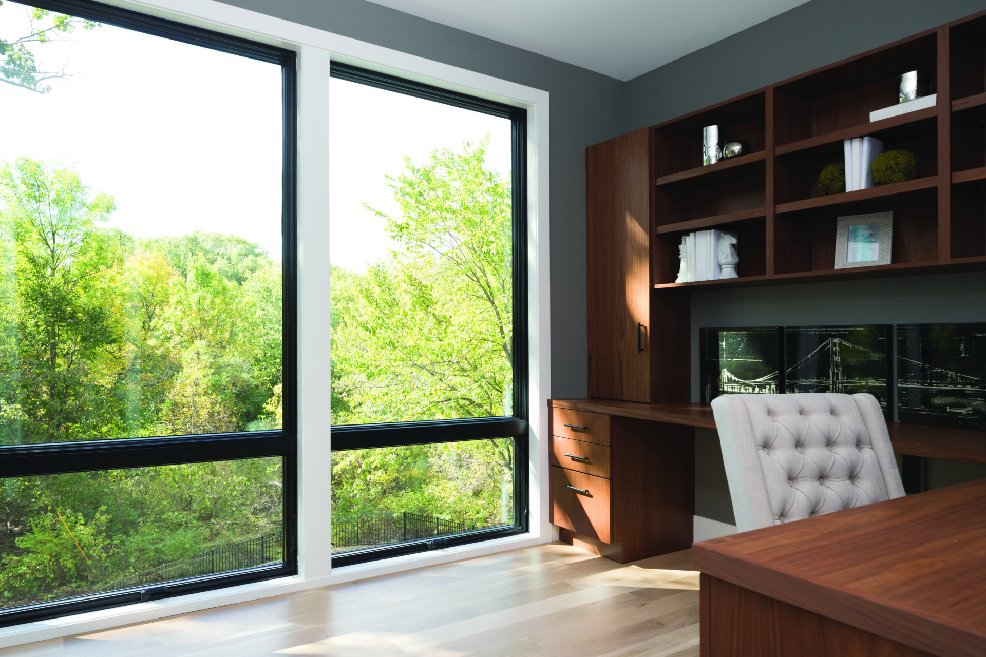 Two large Andersen 400 series awning picture windows overlook a park from the interior of a home office with a dark wood desk and white desk chair.