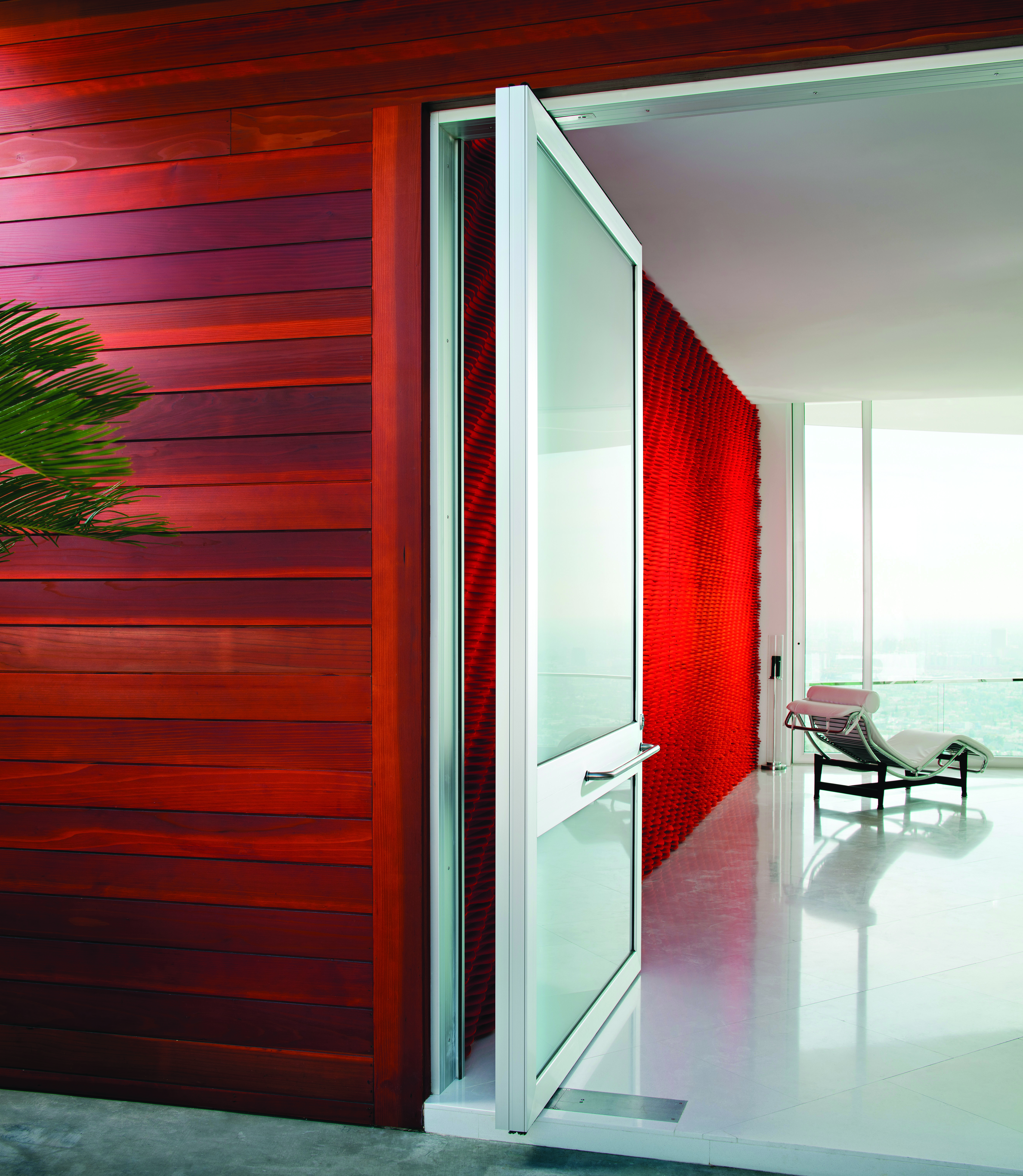 Red paneled interior wall of a minimalist home with a white pivot door open to a room overlooking a floor to ceiling window