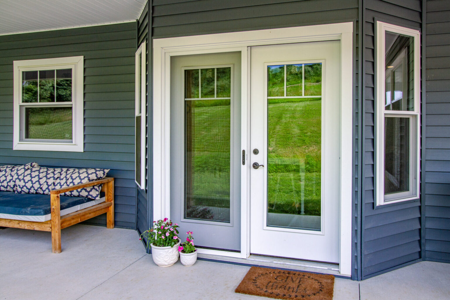 A white-framed hinged patio door with sidelights leads outside from a blue house in Morrisville NC. A brown doormat and two potted plants sit on the concrete porch, and a wooden bench with blue cushions is to the left.