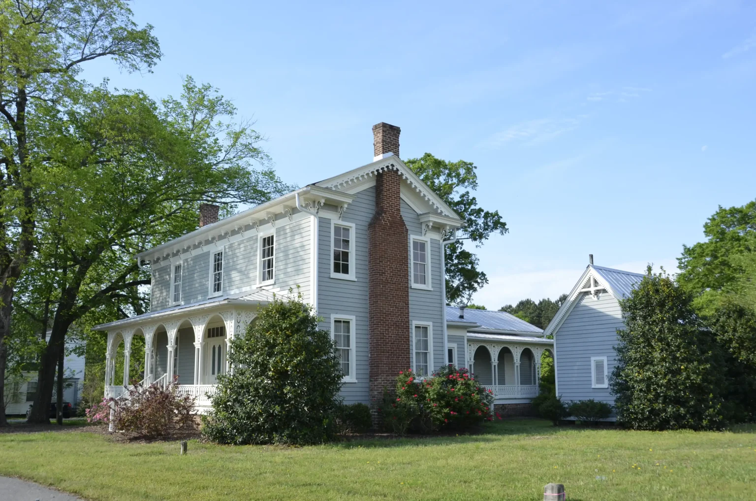 A light blue two-story house with white trim and new window installation, featuring a large brick chimney and wraparound porch, sits on a grassy lawn surrounded by trees and bushes under a clear blue sky.