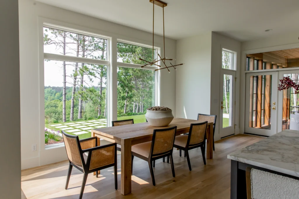 A modern dining room with a wooden table, six chairs, large window installation showcasing a green outdoor view, light wood flooring, and a contemporary chandelier hanging above the table.