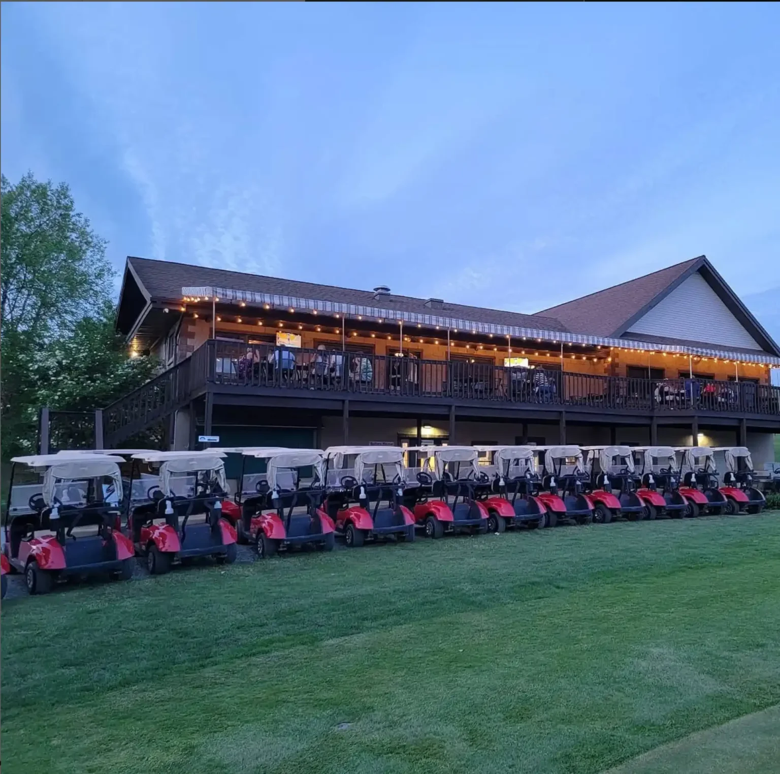 Rows of red and white golf carts are parked on grass in front of a large, two-story building with a balcony and new window installation, where people are seated under string lights in the early evening.