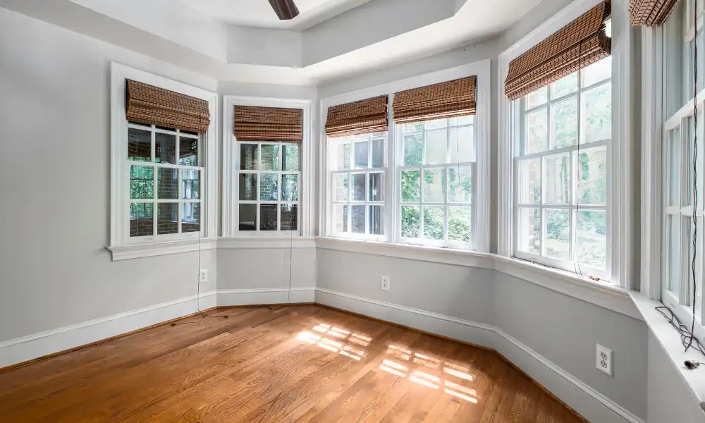 Bright, empty room with light gray walls, hardwood floor, and large bay windows featuring bamboo blinds from a recent window installation, allowing natural light to stream in. Green trees are visible outside the windows.