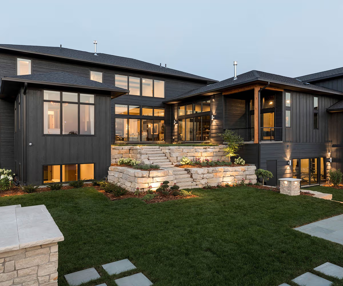 Modern two-story house with dark exterior siding, large windows, stone steps, and landscaped yard. Outdoor lights illuminate the stone features and green lawn in the evening, enhancing curb appeal across our service areas.