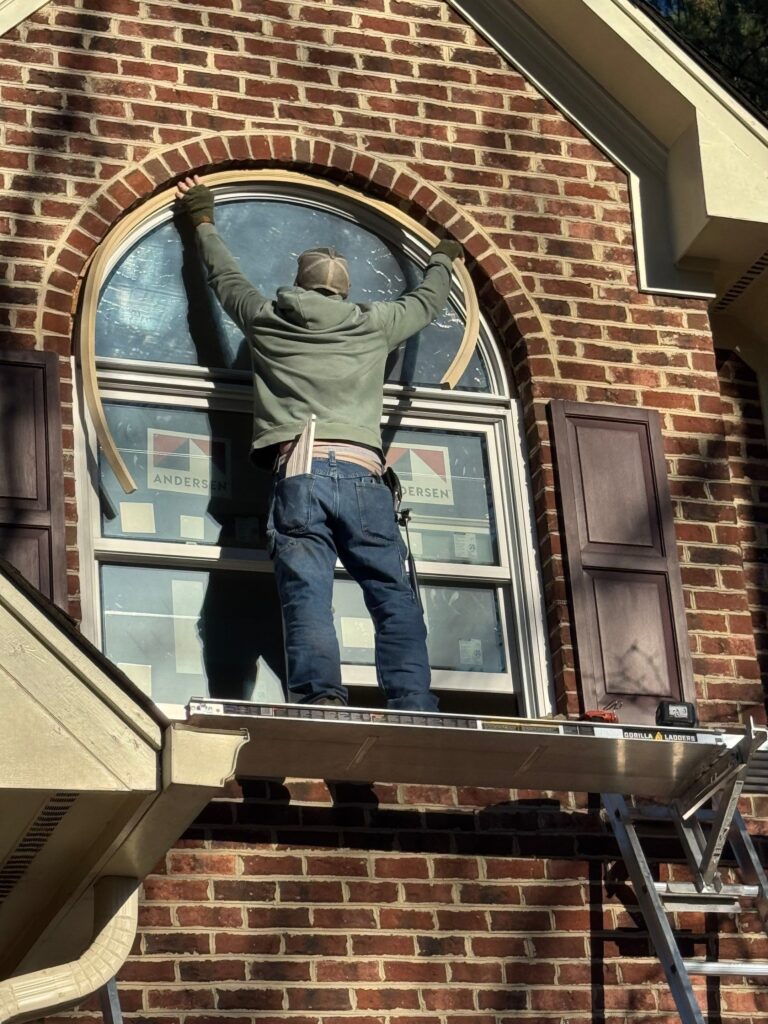 a Window Works Co window installer applies exterior moulding on an arch top Andersen window on the second story of a brick house in Apex, NC