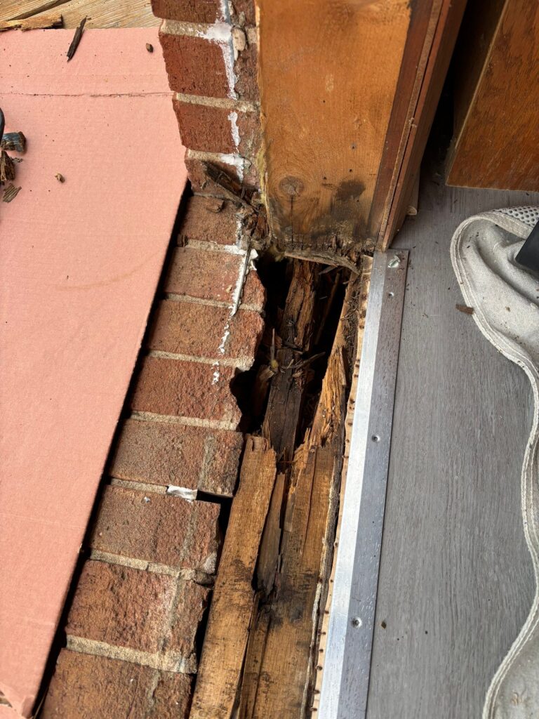 A close-up view of a front door sill is damaged, showing rotting wood and bricks with a large gap exposing decayed material near the threshold. A white shoe and gray floor are visible on the right.
