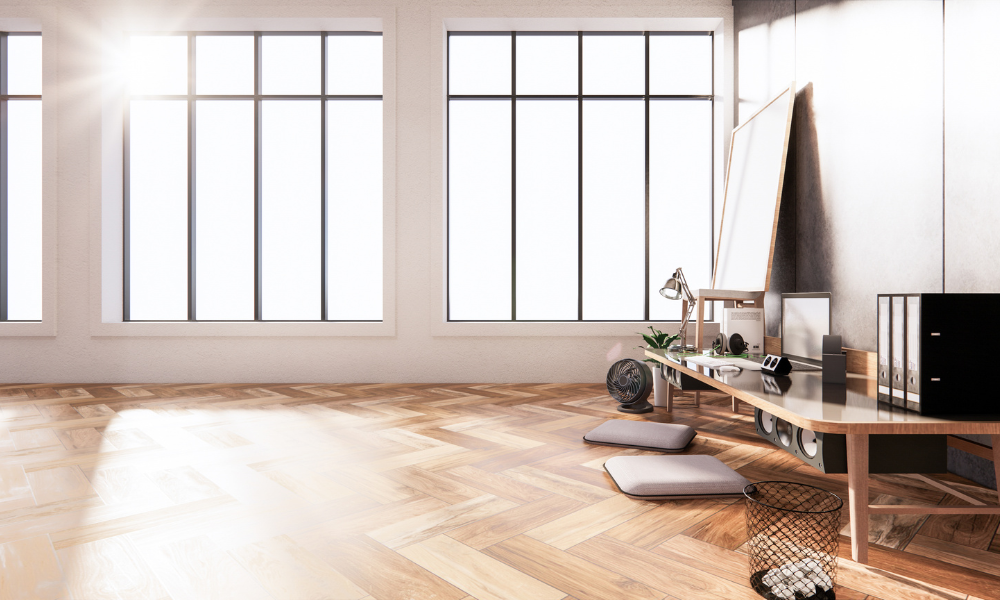 A bright, minimalist room with energy efficient windows, sunlight streaming in, wooden herringbone floor, a low desk with books, plants, lamps, and cushions on the floor. A wire basket sits beside the desk.