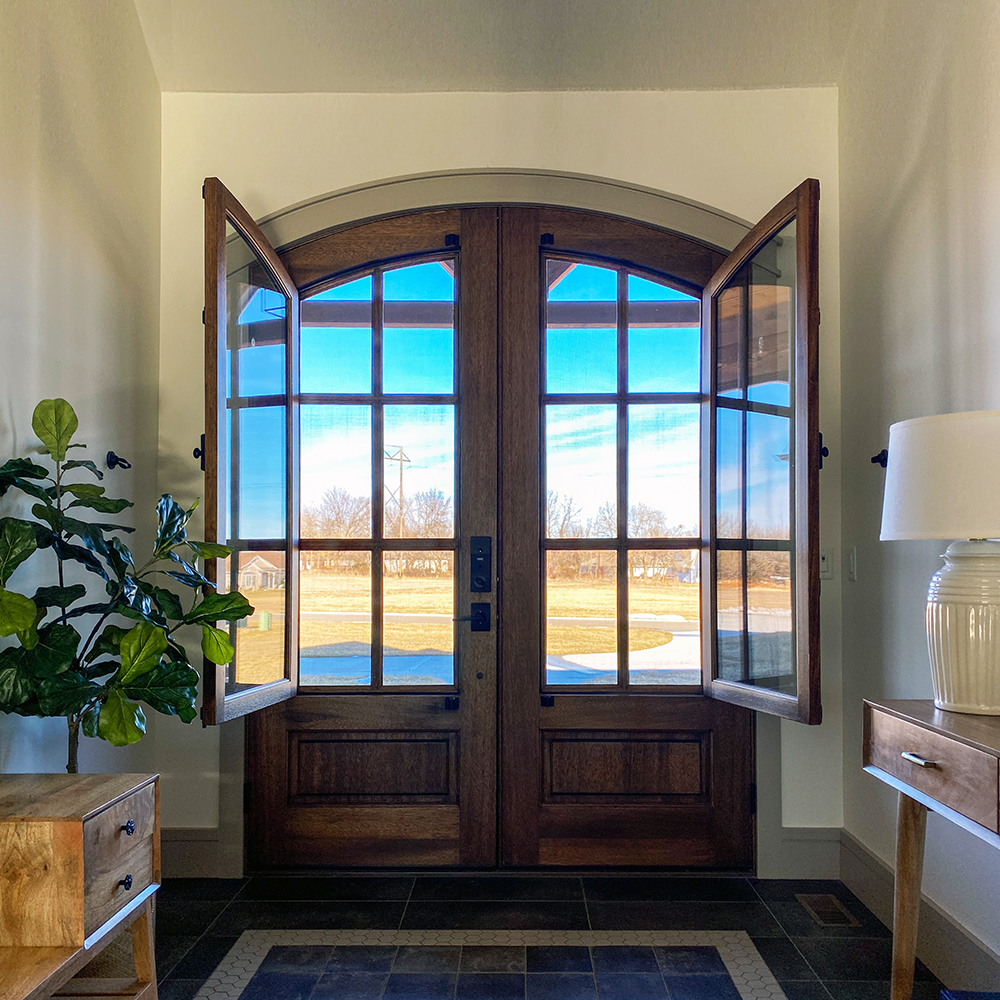 A double wooden arch top door with interior venting lites open in a foyer with a fiddleleaf fig tree. The door is the Breezeport by Raleigh company DSA.