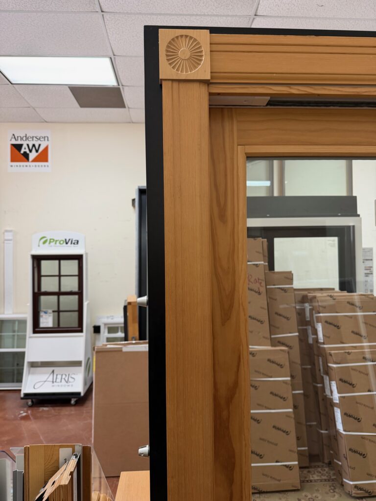 A close-up of a wooden window frame with a decorative carved corner, similar to what is a plinth block on a door, in a showroom. In the background, various boxed products and display windows are visible, along with signs for Andersen, ProVia, and Aeris.