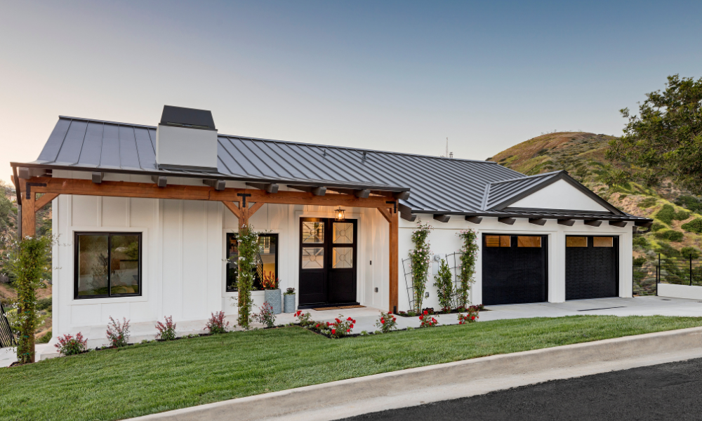 A modern single-story house with white siding, a dark metal roof, large windows from professional window and door installation, and a double garage. A manicured lawn, flower beds, and hills rest in the background under a clear sky.