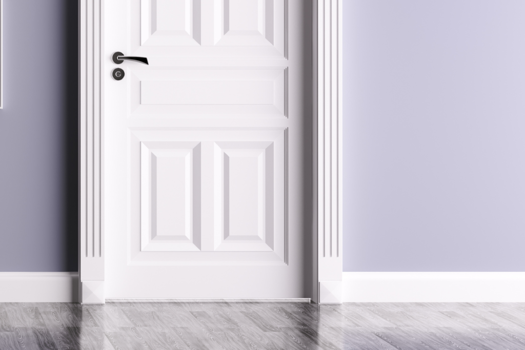 A white, paneled door with a black handle is set in a lavender-colored wall with decorative trim and a plinth block—what is a plinth block on a door? It’s the base piece adding style where trim meets the floor's reflective gray wood planks.