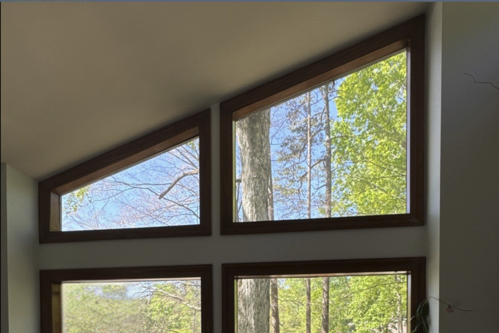 Large triangular and rectangular windows that follow the roofline pitch, with dark wood frames, reveal green trees and a blue sky outside, letting natural light flood the interior space.