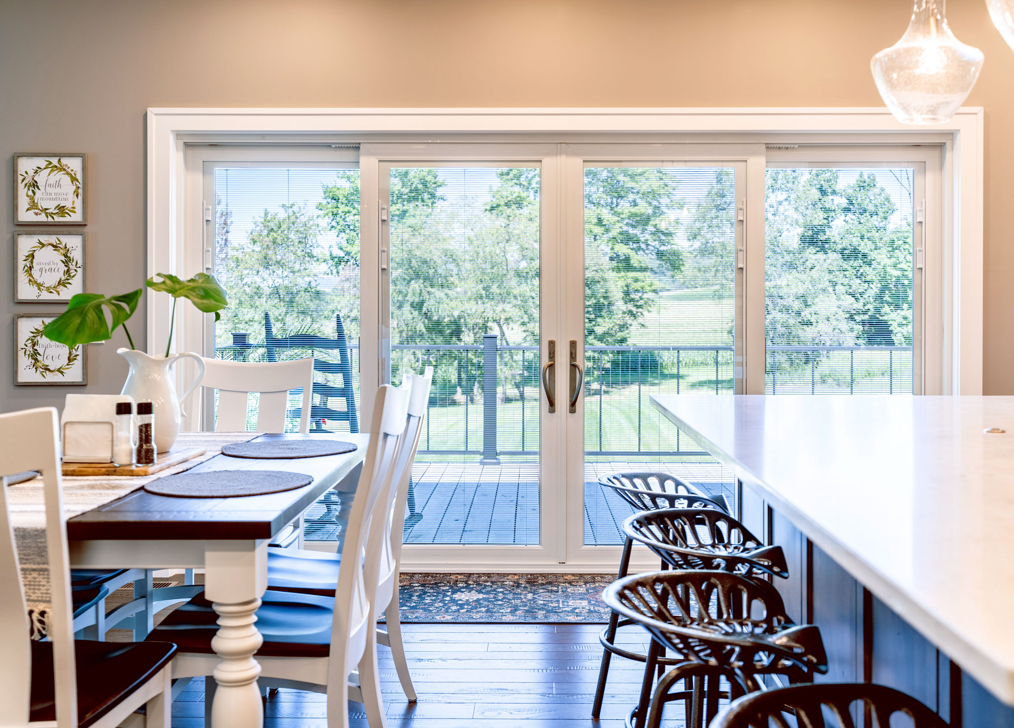 Bright dining area with a table and chairs near a kitchen island with barstools. Large glass doors, featuring professional window and door installation, open to a deck with views of green trees and lawn. Light walls and hardwood floors create an airy vibe.