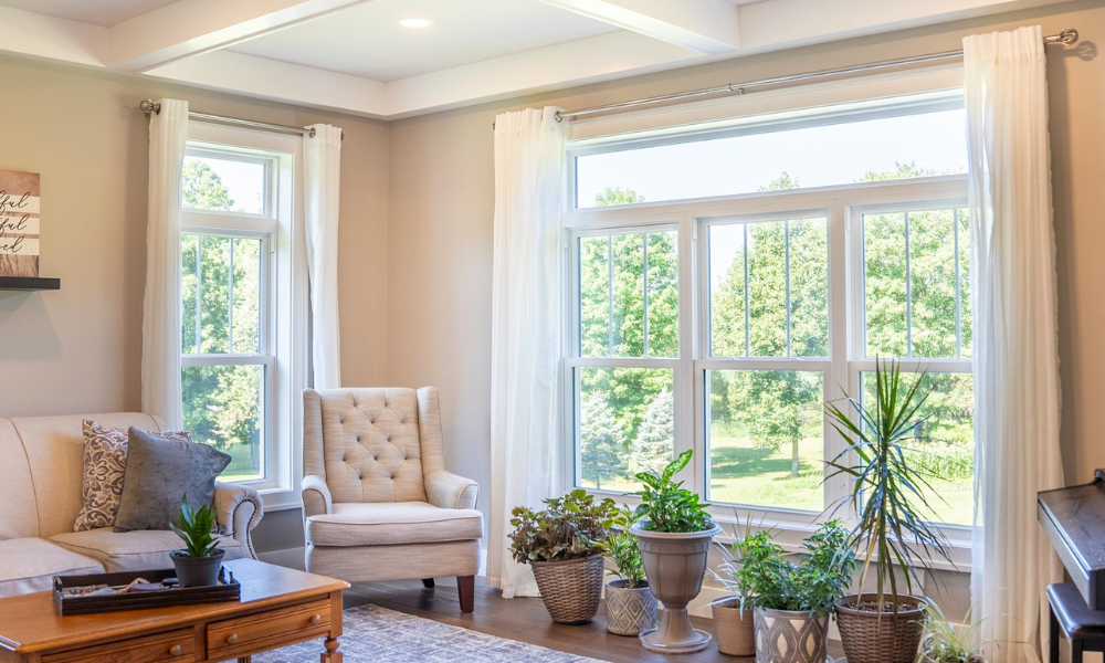 A bright living room with large windows installed by Professional Window and Door Installation lets in natural light. There’s a beige armchair, sofa, wooden coffee table, and potted plants by the windows, with green trees visible outside.