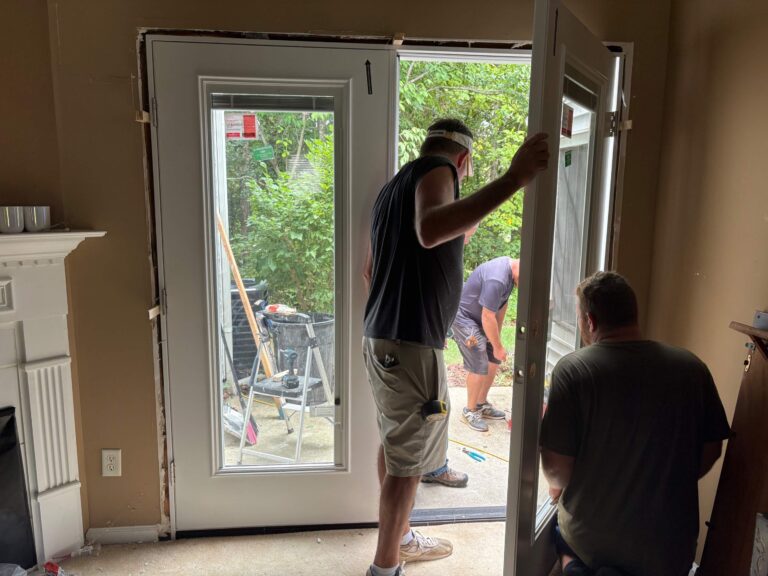 Two men are installing a new white exterior door frame in a residential interior, with one standing and holding the door e...