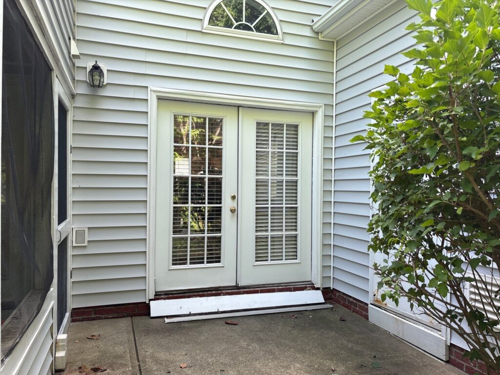 A set of white double french doors with glass panes and closed white blinds is centered on a light gray vinyl-sided wall a...